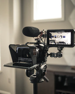 A professional video camera setup includes a large camera mounted on a tripod, equipped with a teleprompter and a monitor. The camera has a microphone attached, and the monitor displays a live feed of the room. The environment appears to be well-lit with natural light coming through a window.