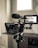 A professional video camera setup includes a large camera mounted on a tripod, equipped with a teleprompter and a monitor. The camera has a microphone attached, and the monitor displays a live feed of the room. The environment appears to be well-lit with natural light coming through a window.