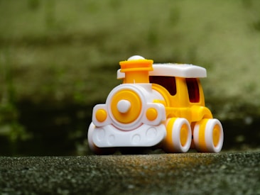 A small, colorful toy train with a bright yellow and white color scheme sits on a textured surface. The locomotive features smooth, rounded shapes and is set against a blurred green background.