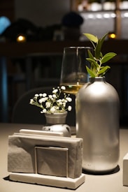 A modern and minimalistic table setting featuring a sleek silver vase with green leaves next to a smaller glass container holding delicate white flowers. In the background, a wine glass half-filled with white wine is visible, creating an elegant and sophisticated atmosphere.