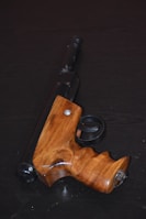 A wooden-handled harpoon gun is displayed on a dark surface. The handle is smoothly crafted with a natural wood finish, contrasting with the dark metal barrel and trigger mechanism.