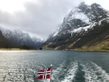 A fjord with steep snow-capped mountains on either side under a cloudy sky. A flagpole mounted on a vessel displays the Norwegian flag over the water.