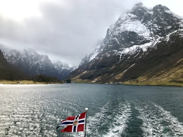 A fjord with steep snow-capped mountains on either side under a cloudy sky. A flagpole mounted on a vessel displays the Norwegian flag over the water.