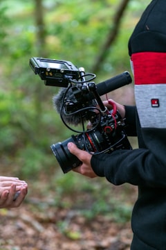 A person is holding a professional camera setup that includes a microphone and a monitor, set against a natural, green outdoor background. The camera appears to be a DSLR or similar type designed for high-quality video recording.