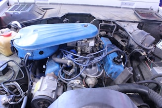 A detailed view of a car engine with various components visible, including a blue air filter cover, wires, hoses, and metal parts. The engine is set within an older vehicle's engine bay, with signs of wear and dirt in the surroundings.