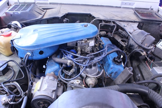 A detailed view of a car engine with various components visible, including a blue air filter cover, wires, hoses, and metal parts. The engine is set within an older vehicle's engine bay, with signs of wear and dirt in the surroundings.