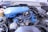 A detailed view of a car engine with various components visible, including a blue air filter cover, wires, hoses, and metal parts. The engine is set within an older vehicle's engine bay, with signs of wear and dirt in the surroundings.