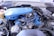 A detailed view of a car engine with various components visible, including a blue air filter cover, wires, hoses, and metal parts. The engine is set within an older vehicle's engine bay, with signs of wear and dirt in the surroundings.