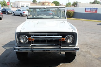 A vintage white SUV is parked on a paved area, with several buildings in the background. The vehicle has a classic design, featuring a distinct front grille and round headlights. The area appears to be a car service or parking lot, with other cars visible in the vicinity.