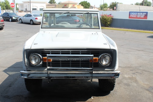 A vintage white SUV is parked on a paved area, with several buildings in the background. The vehicle has a classic design, featuring a distinct front grille and round headlights. The area appears to be a car service or parking lot, with other cars visible in the vicinity.
