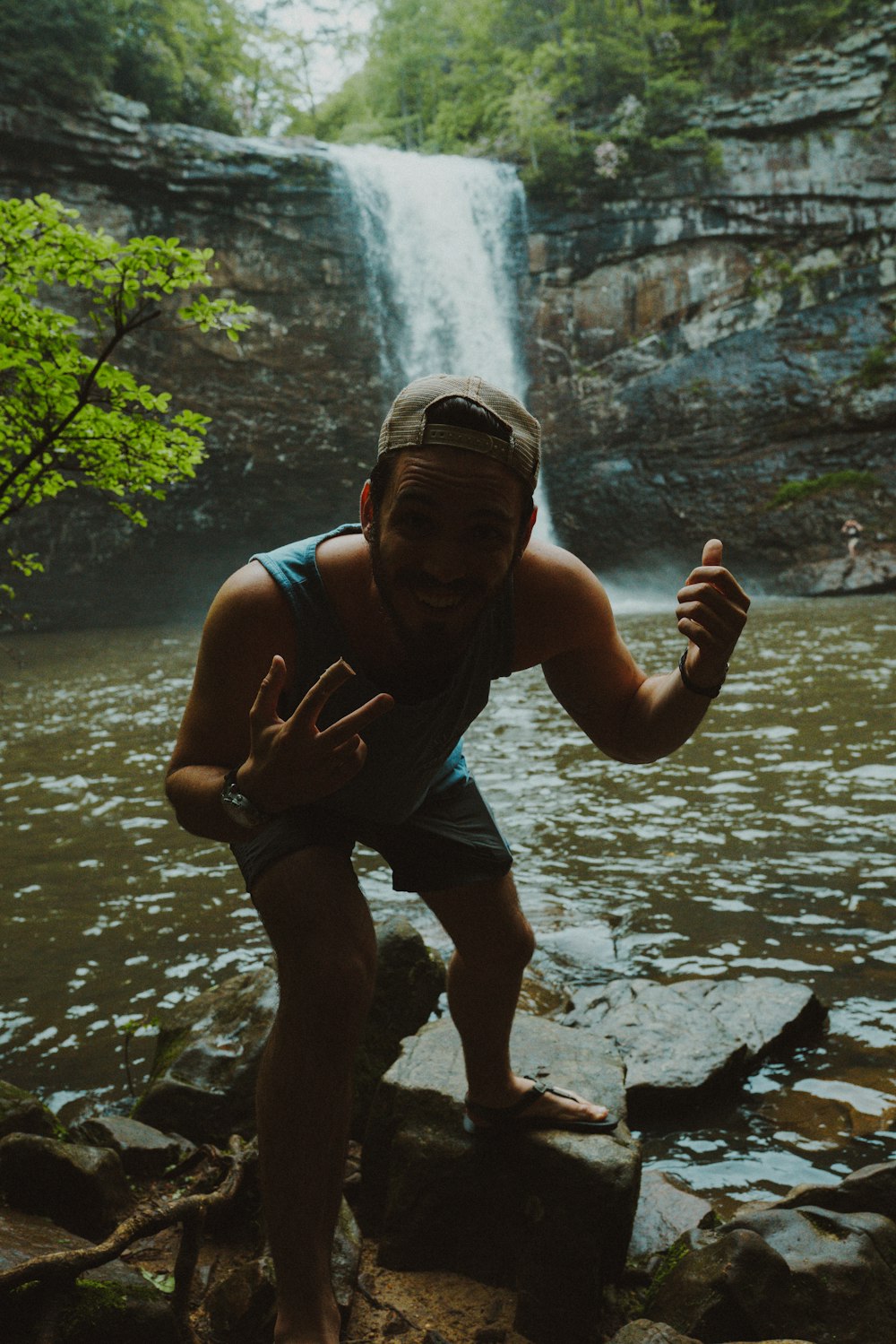 man in blue tank top and green shorts sitting on rock near river during daytime