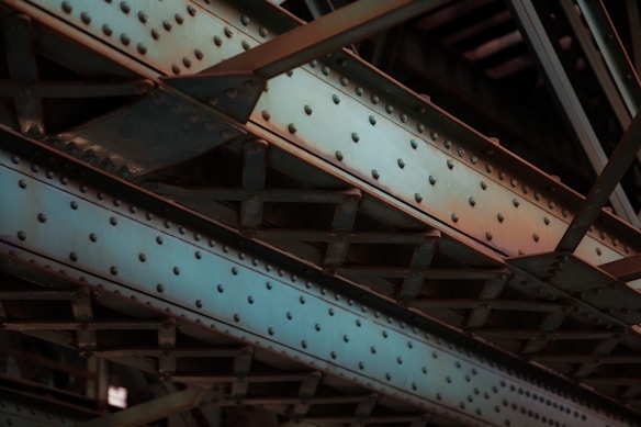 A close-up view of industrial steel girders and beams with a riveted design, predominantly colored in steel blue and rusty orange shades. The intersecting lines and angles create a complex structure, showcasing the metallic texture and utilitarian design.