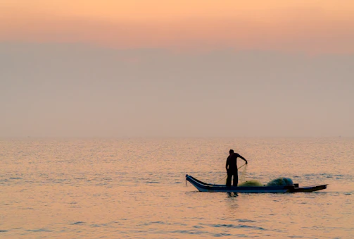 A barefoot local fisherman casting his net with the turquoise sea and rocky cliffs of Nusa Penida in the background during golden hour.