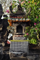 An intricately carved stone shrine adorned with colorful fabric and surrounded by lush greenery and pink flowers. The structure features traditional architectural details and patterns, conveying a cultural and artistic aesthetic.
