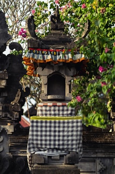 An intricately carved stone shrine adorned with colorful fabric and surrounded by lush greenery and pink flowers. The structure features traditional architectural details and patterns, conveying a cultural and artistic aesthetic.