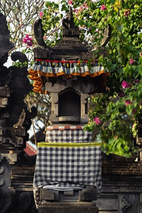 An intricately carved stone shrine adorned with colorful fabric and surrounded by lush greenery and pink flowers. The structure features traditional architectural details and patterns, conveying a cultural and artistic aesthetic.