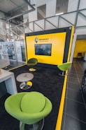 A modern showroom interior with a bright yellow Renault display wall featuring a screen. Green, stylish chairs with metallic bases are placed in a seating area on a dark carpet with yellow borders. Glass panels and a grey staircase with metal railings are visible in the background, creating an industrial yet polished atmosphere.