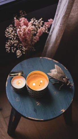 A rustic wooden stool with a weathered teal surface holds two lit candles in decorative holders. A large metallic insect sculpture adds an artistic touch. In the background, dried flowers with pink and cream hues stand beside a sheer beige curtain.