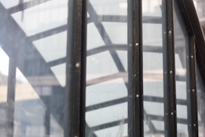 The image features a close-up view of a glass structure with metal frames. The geometric pattern formed by the metal frames creates a modern architectural element, reflected onto the glass panels. The background reveals a blurred cityscape, adding depth to the composition.