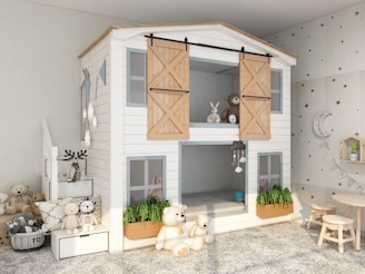 A cozy and well-decorated children's play area featuring a small, house-shaped bunk bed with white wooden planking and light wooden sliding doors on the upper level. The bottom level has open windows with potted greenery below them. Soft toys, including teddy bears and a rabbit, are placed throughout the room. Some toys are on a small white staircase to the left, which leads to the upper level of the bunk bed. The room also has a small wooden table and two stools on the right, along with decorative items like string lights, a cloud-shaped wall shelf, and star decals on the walls.