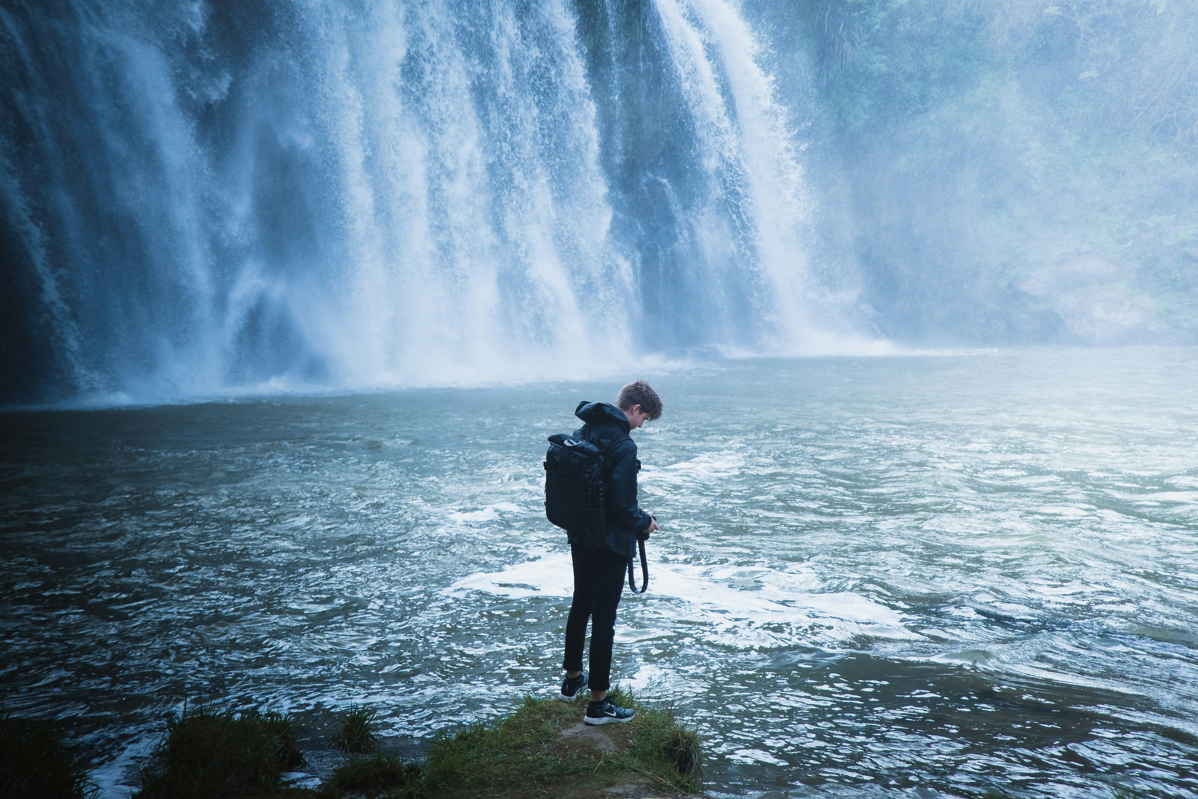 man in black jacket standing on green grass near water falls during daytime