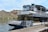 A large tour boat called 'Desert Belle' is docked at a lake pier. The boat has two levels with white railings and numerous white chairs, indicating seating for passengers. The backdrop features a hilly terrain with sparse vegetation typical of a desert landscape under a partly cloudy sky.