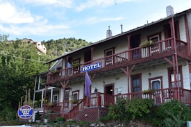 A rustic two-story hotel building with wooden railings and balconies, surrounded by greenery and set against a hillside. The hotel sign is prominently displayed, and potted plants are placed along the balconies.