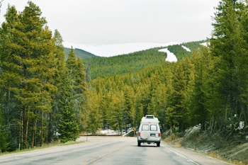 A van travels along a paved road surrounded by dense evergreen forests. Snow-capped mountains rise in the background, under an overcast sky. Road signs indicate a curve ahead, and the scene conveys a sense of wanderlust and adventure.