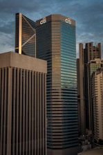 A cityscape featuring a cluster of modern skyscrapers with a large building prominently displaying the Citi logo. The architecture is sleek and contemporary, with glass and concrete facades reflecting ambient light. The sky is overcast, providing a moody backdrop to the urban setting.