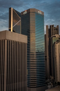 A cityscape featuring a cluster of modern skyscrapers with a large building prominently displaying the Citi logo. The architecture is sleek and contemporary, with glass and concrete facades reflecting ambient light. The sky is overcast, providing a moody backdrop to the urban setting.