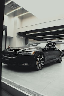 A sleek, black luxury car is parked indoors under modern lighting. The reflective surface of the car highlights its polished finish and sharp design lines. The setting appears to be a sophisticated showroom with ceiling lights casting a soft glow.
