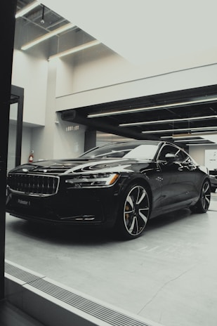 A sleek, black luxury car is parked indoors under modern lighting. The reflective surface of the car highlights its polished finish and sharp design lines. The setting appears to be a sophisticated showroom with ceiling lights casting a soft glow.