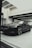 A sleek, black luxury car is parked indoors under modern lighting. The reflective surface of the car highlights its polished finish and sharp design lines. The setting appears to be a sophisticated showroom with ceiling lights casting a soft glow.