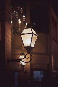 An ornate wall-mounted lantern casts a soft glow in the dim lighting of a rustic interior. Behind it, a chandelier with multiple lights is visible, adding to the warm ambiance. The background features brick walls and hints of wooden elements, enhancing the cozy and vintage atmosphere.
