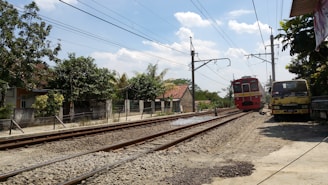 A red commuter train moves along railroad tracks in a rural area, passing by houses and trees. Overhead power lines run above the tracks, and a truck is parked nearby on the side of the road. Clear blue skies with some clouds complete the scene.