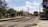 A red commuter train moves along railroad tracks in a rural area, passing by houses and trees. Overhead power lines run above the tracks, and a truck is parked nearby on the side of the road. Clear blue skies with some clouds complete the scene.