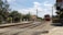 A red commuter train moves along railroad tracks in a rural area, passing by houses and trees. Overhead power lines run above the tracks, and a truck is parked nearby on the side of the road. Clear blue skies with some clouds complete the scene.