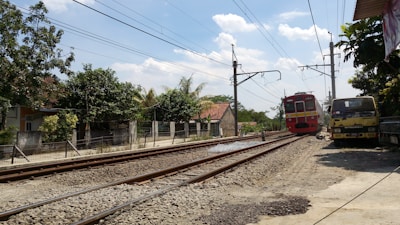 A red commuter train moves along railroad tracks in a rural area, passing by houses and trees. Overhead power lines run above the tracks, and a truck is parked nearby on the side of the road. Clear blue skies with some clouds complete the scene.