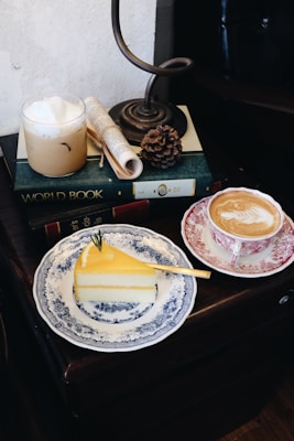 A cozy setup includes a slice of yellow cake on a decorative blue and white plate with a small green garnish. Nearby, there is a cup of latte with foam art in a red and white patterned cup. An iced coffee sits on a stack of books labeled 'World Book', '1', and '2'. A small rolled-up newspaper and a pinecone are also present, all under an antique-style lamp.