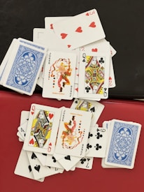 A collection of playing cards is spread out haphazardly on a surface. The cards include various face cards, jokers, and number cards from different suits, with some cards face down showing a blue patterned back. The visible cards display vibrant illustrations with a mix of red, black, and yellow elements, against a predominantly white background.