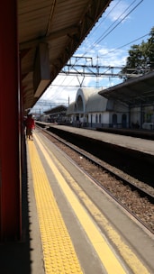 A train station platform featuring a covered walkway with metal roofing. The platform is lined with yellow tactile paving for the visually impaired. Overhead electric lines are visible, along with tracks running alongside the platform. A few people are walking along the platform, with a station building visible in the background under a partly cloudy blue sky.