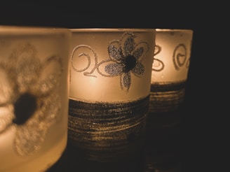 Three decorative candle holders with floral patterns and swirling designs sit in a dimly lit environment. The glow of the candlelight creates a warm and intimate ambiance.
