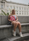 A person is sitting on a stone bench holding a red book or magazine that says 'WESTMONT'. The background features a classical building with large windows and some greenery.