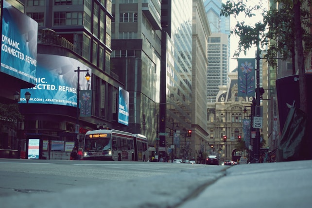 A bustling urban street scene with tall buildings lining both sides. An advertisement with the words 'dynamic, connected, refined' is prominently displayed on a digital billboard. A city bus with a '48 Front-Market' route sign is moving along the road. The background features the architectural details of an ornate historic building. Trees and traffic lights are visible, and pedestrian sidewalks are present.