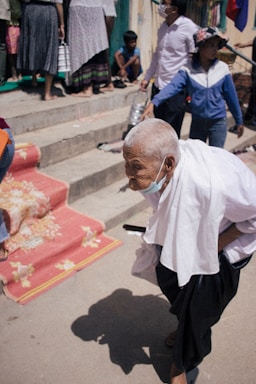 An elderly person wearing a white shirt and black pants is walking with assistance, holding a cane. The person is wearing a face mask and appears to be in an outdoor setting with others around. There are stepping stairs in the background and a red carpet with floral patterns leading up the steps. Other people, some seated and some standing, can also be seen.