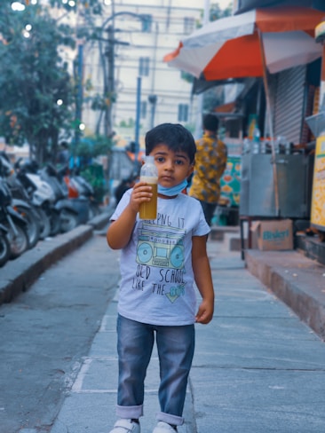 A young child stands on a sidewalk holding a bottle containing a greenish beverage. The child is wearing a grey T-shirt with a graphic of a boombox and text, along with folded jeans and white shoes. The background shows a busy street scene with motorcycles parked along the edge, and a person wearing colorful clothing walking past a street vendor's stall with an orange and white umbrella.