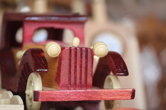 A wooden toy car with a vintage design, featuring prominent front grille and round headlights. Made from polished wood, it displays a rich, dark red color with a contrasting natural wood hue on the side panels. The focus is on the front of the car, with blurred background elements, giving a sense of depth.