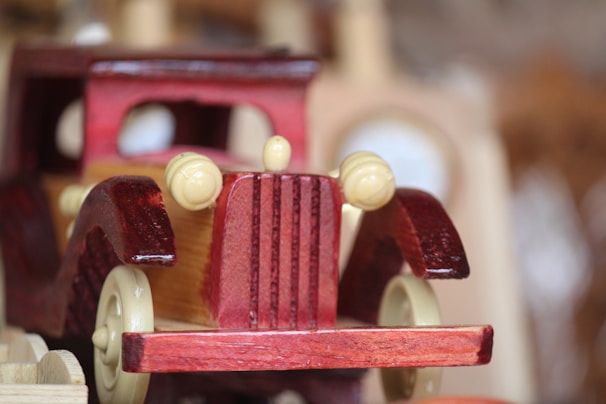 A wooden toy car with a vintage design, featuring prominent front grille and round headlights. Made from polished wood, it displays a rich, dark red color with a contrasting natural wood hue on the side panels. The focus is on the front of the car, with blurred background elements, giving a sense of depth.