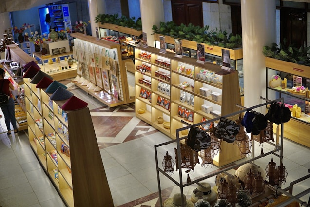 A well-organized gift shop with wooden shelves displaying a variety of products such as hats, bags, souvenirs, and other items. The shop features bright lighting and decorative plants on top of the shelves. The layout is neat, with sections for specific types of merchandise.