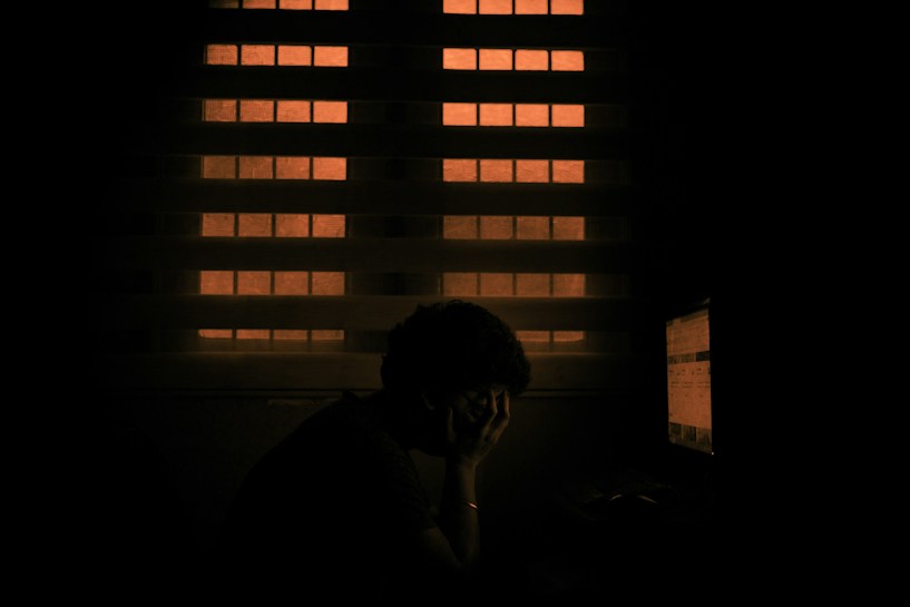A dimly lit room features a person sitting in front of a computer screen with a hand resting on their face, conveying a sense of fatigue or contemplation. The background is illuminated by a window with a warm orange light.